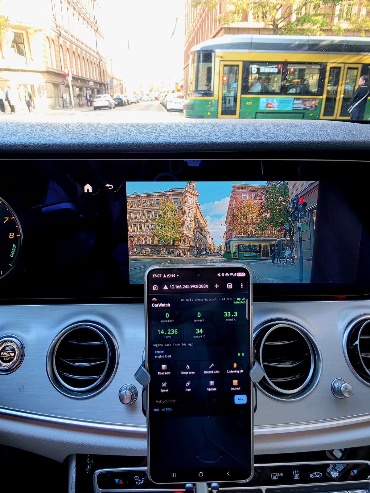 Phone in a Mercedes showing CarWatch live: 0 km/h, 0 rpm, 33.3 percent hybrid, 14.2 V, coolant 34 C, parked on a Helsinki street with a tram