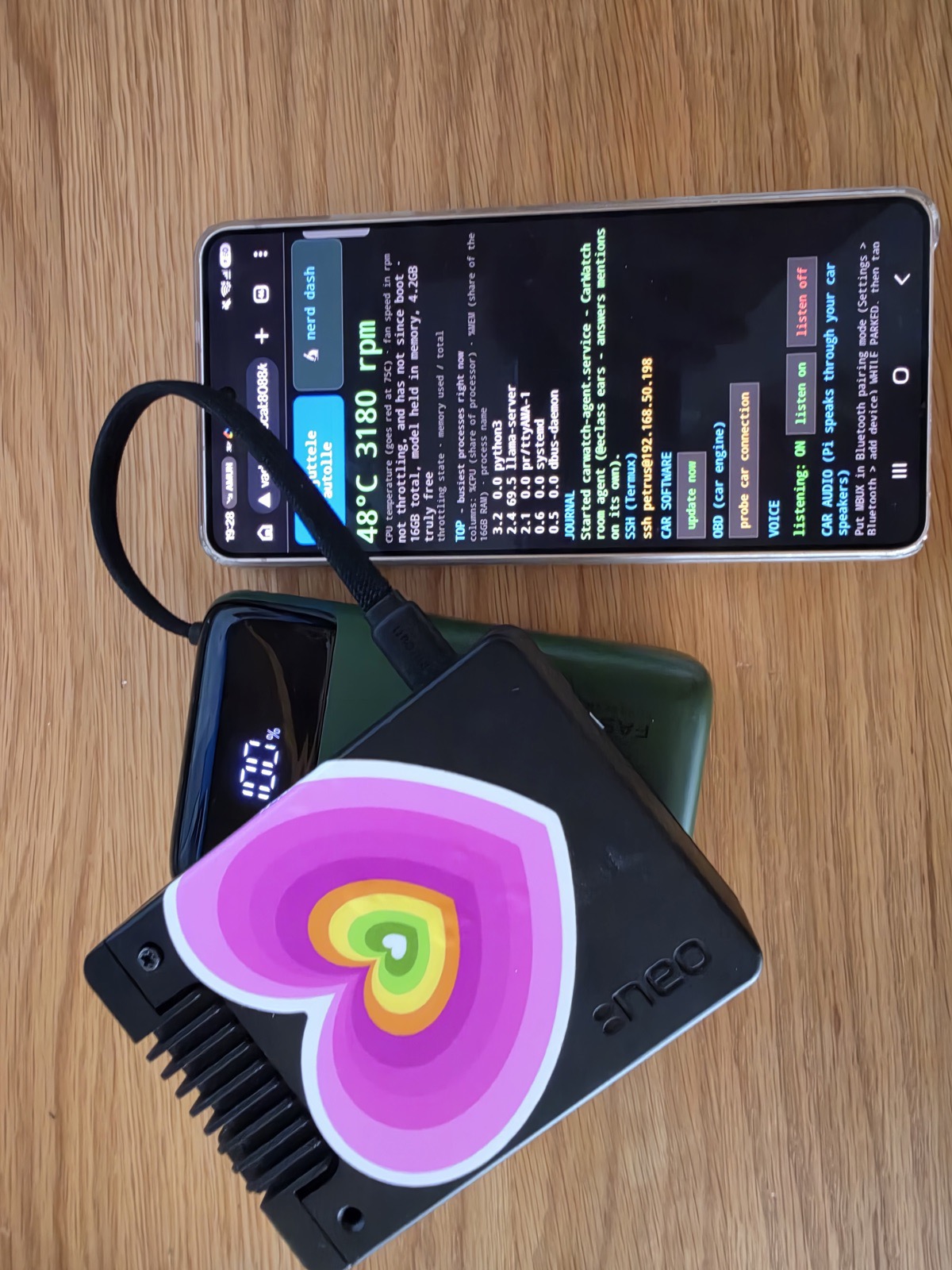 The CarWatch rig: a Raspberry Pi in a black heatsink case with a rainbow heart sticker, a power bank, and a phone showing the live dashboard